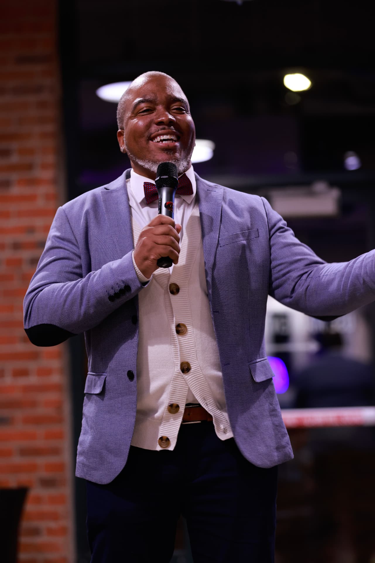 Jay.R Fogle smiling with a microphone in a grey blazer and bow tie